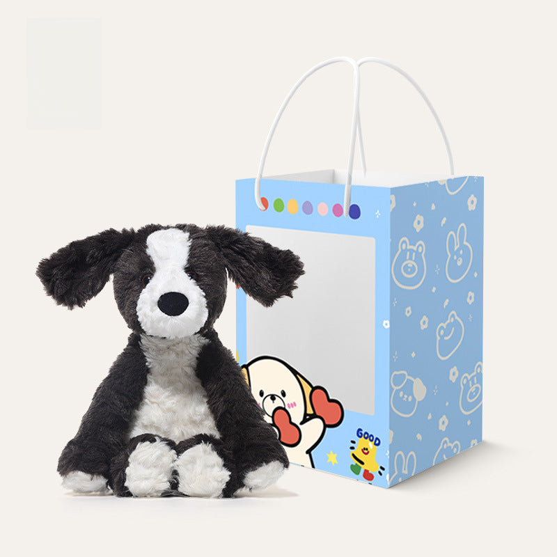 The 24cm JOOKI Black and White Puppy Plush Toy, a soft stuffed dog with floppy ears, sits in front of a blue gift bag adorned with cartoon animal faces and colorful dots.