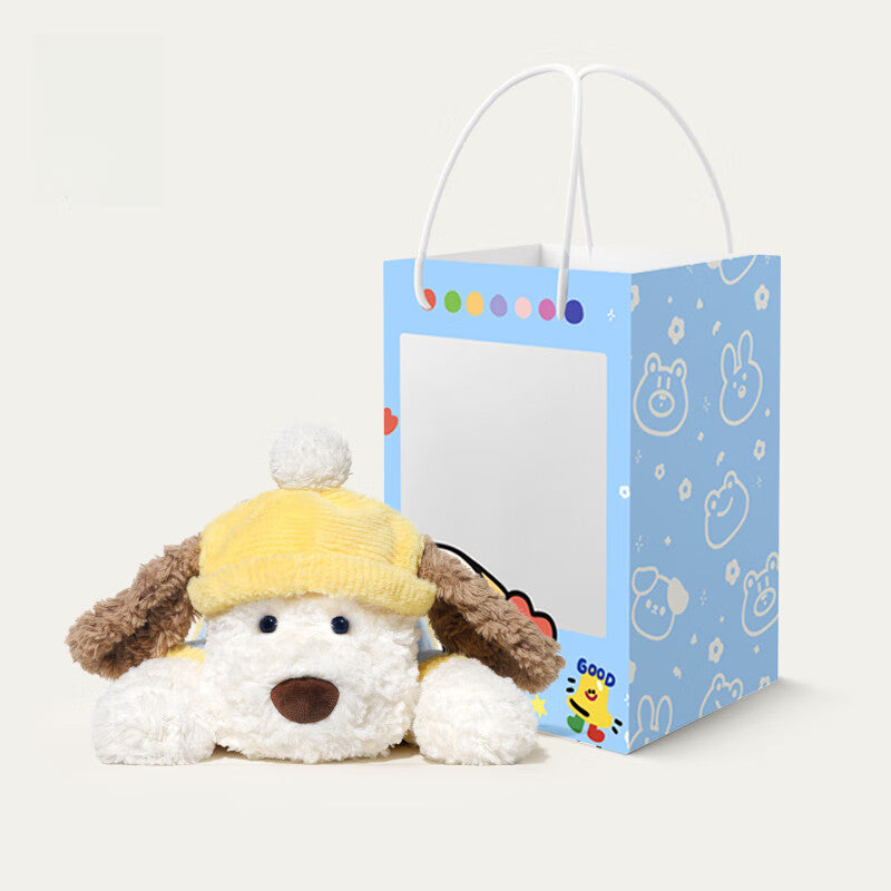 The JOOKI 44cm Lying Dog Plush Toy with Yellow Hat sits next to a blue gift bag with animal faces and dots—a cute, soft stuffed puppy for kids and decor, perfect for plushie fans.