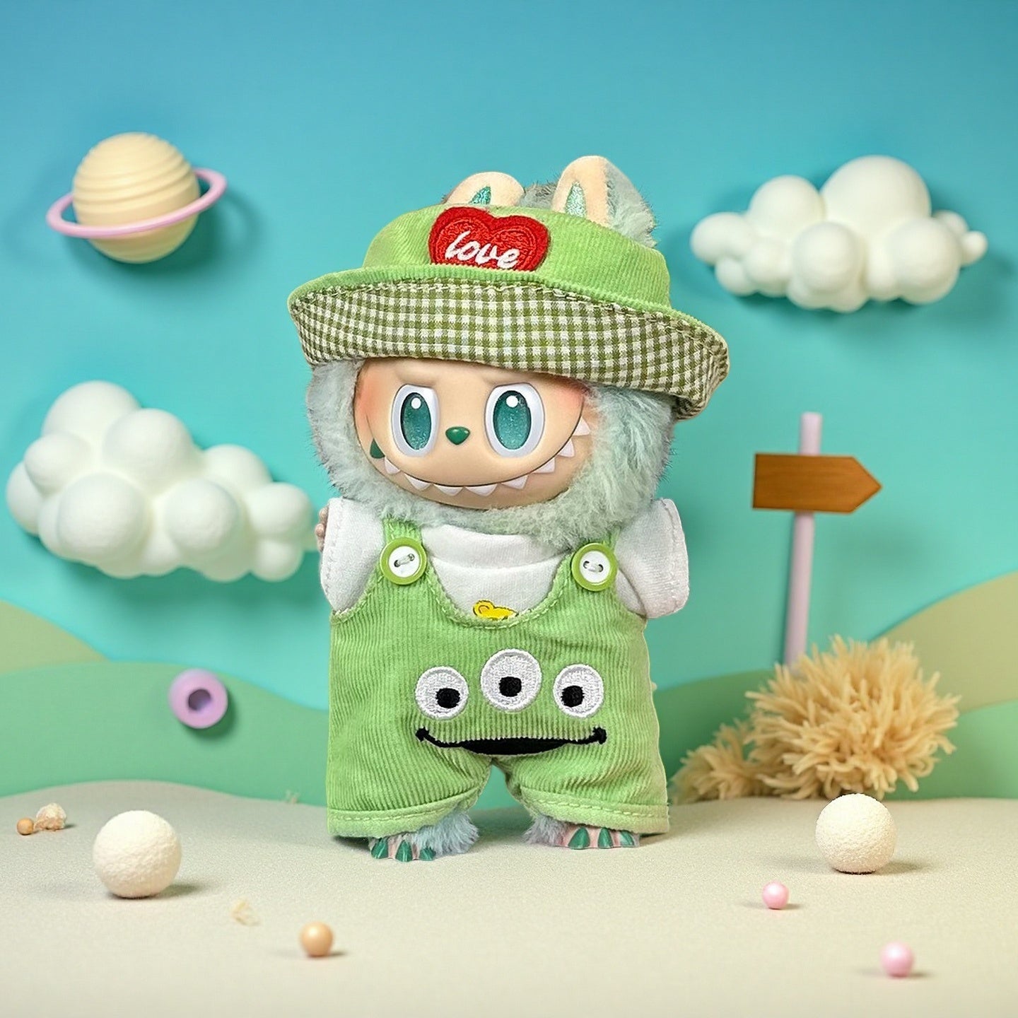 The ZoDoll Labubu Green Corduroy Monster Overalls and Gingham Bucket Hat Set features bunny ear accents, shown on a plush doll posed against a pastel backdrop with clouds and planets—ideal for dressing up your Labubu.