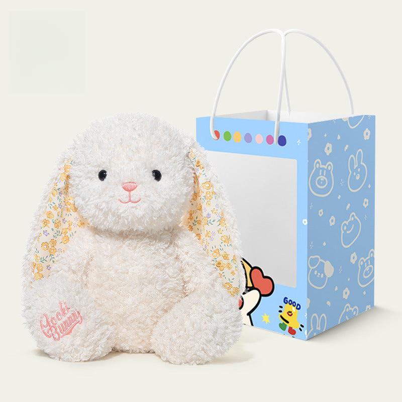 The JOOKI 38cm White Bunny Plush Toy, featuring floral ears and an embroidered paw, sits next to a blue cartoon-themed gift bag, making a perfect Easter gift.