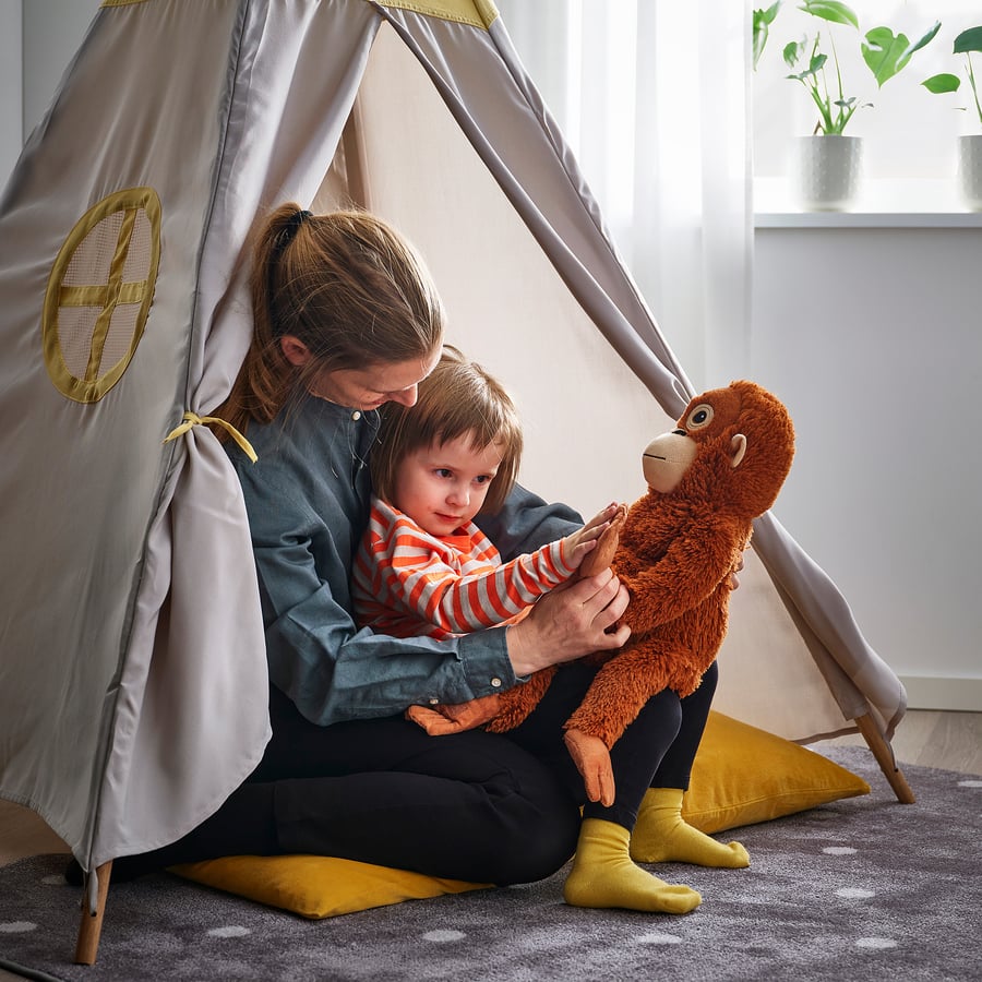 An adult sits with a child inside a fabric play tent. The child, seated on the adult’s lap, holds a ZoDoll Giant Punch Monkey Plush Toy—Viral Djungelskog Orangutan. Cozy pillows and plants appear in the background.