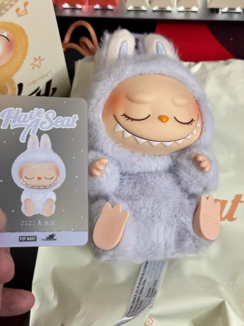 A Labubu “Zizi” plush keychain from POP MART’s Have a Seat Series, in a fuzzy gray bunny suit with closed eyes and a wide smile, rests on a bag beside its matching collectible card.