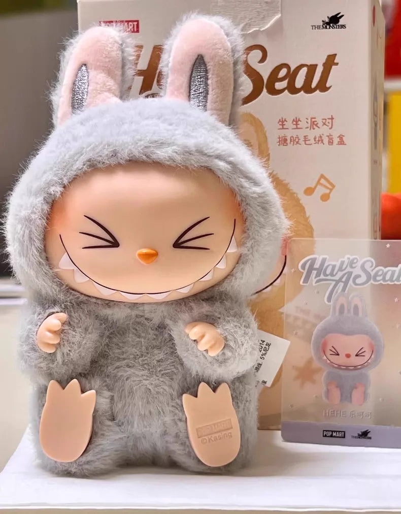 The Labubu “Hehe” Plush Keychain – Have a Seat Series Gray Bunny by POP MART sits before its box and matching card, both showcasing the same Pop Mart The Monsters character design.
