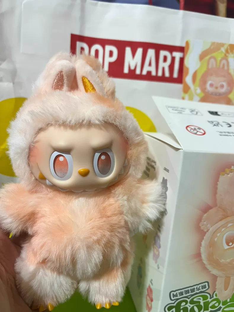 The Labubu “Happiness” Plush Keychain – The Monsters “Big Into Energy” Series Peach Bunny Bag Charm by POP MART is held in front of its matching box and a POP MART shopping bag, perfect for any fan’s collection.