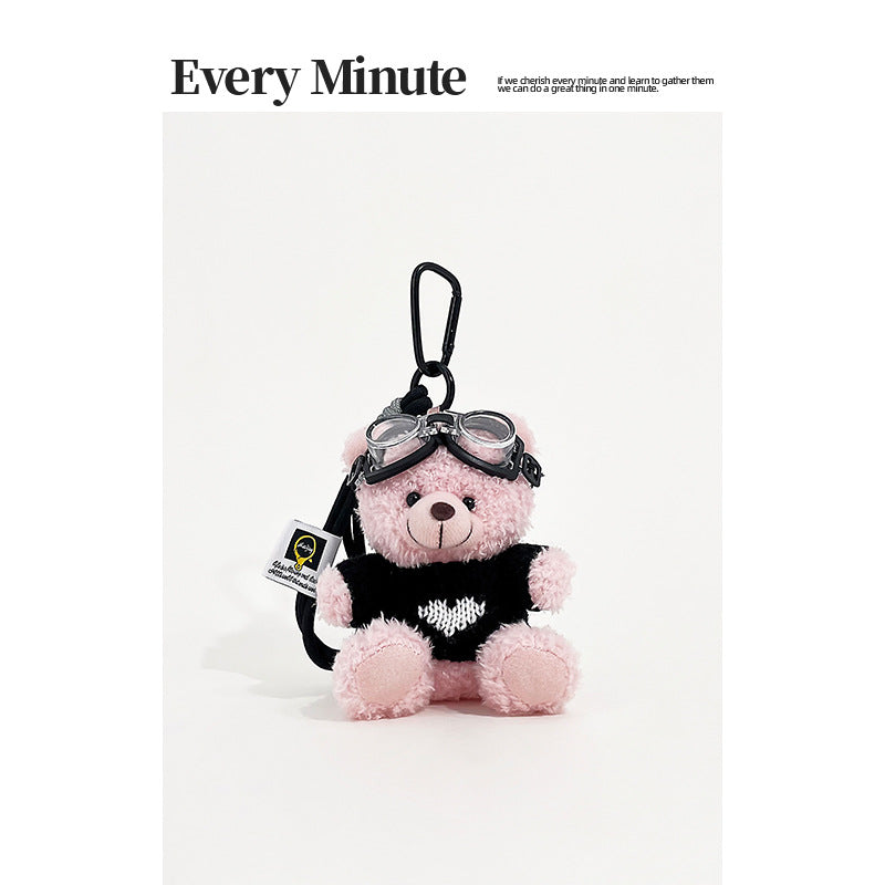 Pink bear plush keychain charm wearing aviator goggles and a black sweater with white heart motif and black carabiner clip