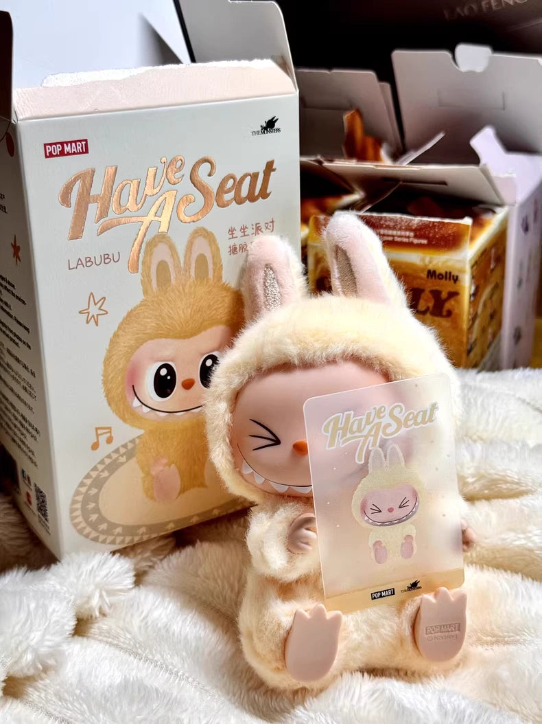 The Labubu “Sisi” Plush Keychain – Have a Seat Series Yellow Bunny by POP MART The Monsters sits with its packaging and a matching card—a must-have collectible for POP MART fans.