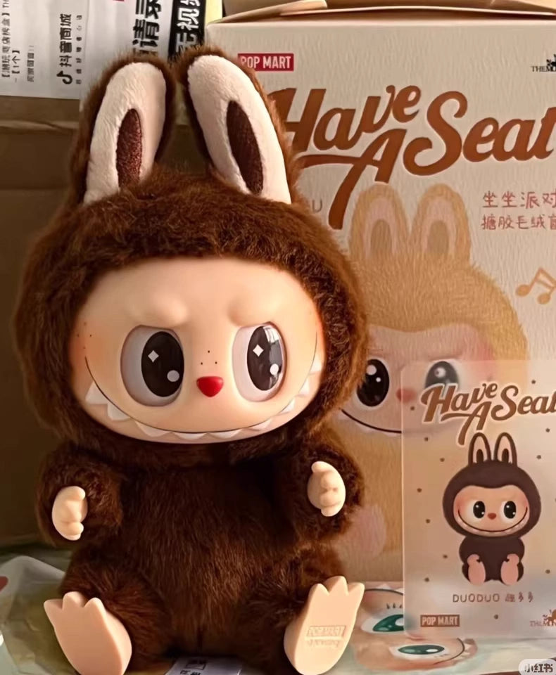 A round-eyed, doll-like figure in a brown bunny suit stars as the Labubu “Duoduo” Plush Keychain – Have a Seat Series Brown Freckled Bunny by POP MART The Monsters, shown with its matching box and card.