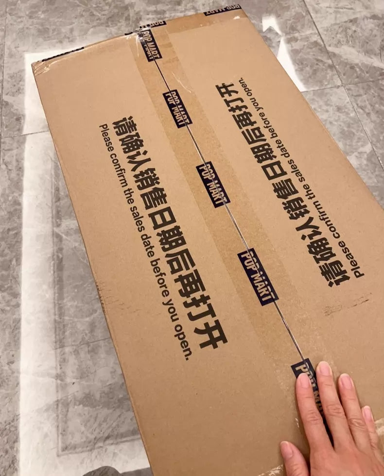 A hand rests on a large sealed POP MART box marked "Please confirm the sales date before you open." It likely contains the Labubu Angel in Clouds 58cm Designer Plush Doll – THE MONSTERS Premium Limited Edition.
