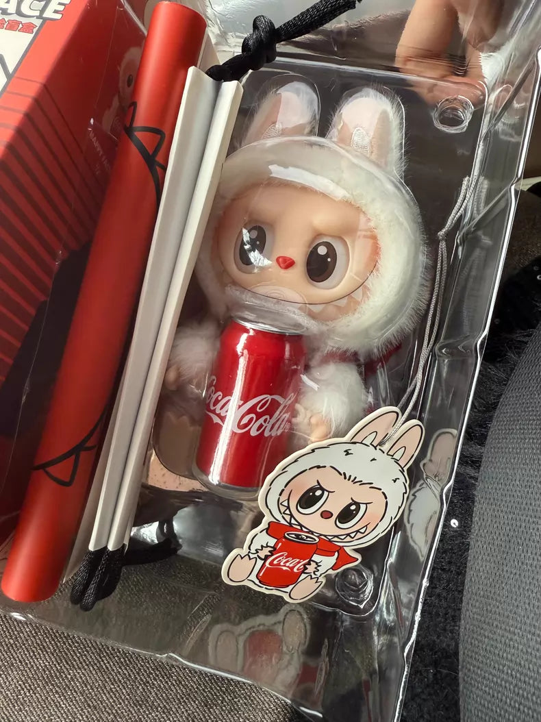 The POP MART Labubu Coca‑Cola Happy Factor Plush Keychain – White Bunny Vinyl Collectible is displayed in a plastic case with a matching bunny sticker and rolled papers—ideal for collectors of Coca‑Cola merchandise.