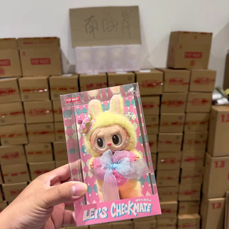 A hand holds a boxed POP MART Labubu Let's Checkmate Series – Pink Heart Bunny Plush Vinyl Doll in front of stacks of cardboard boxes.