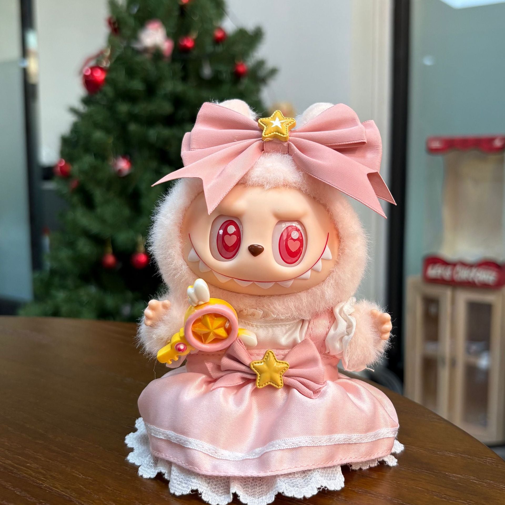 A ZoDoll Labubu Pink Magical Girl plush doll, dressed in a satin lace dress with bows, stars, and holding a star wand, sits on a table with a decorated Christmas tree in the background.