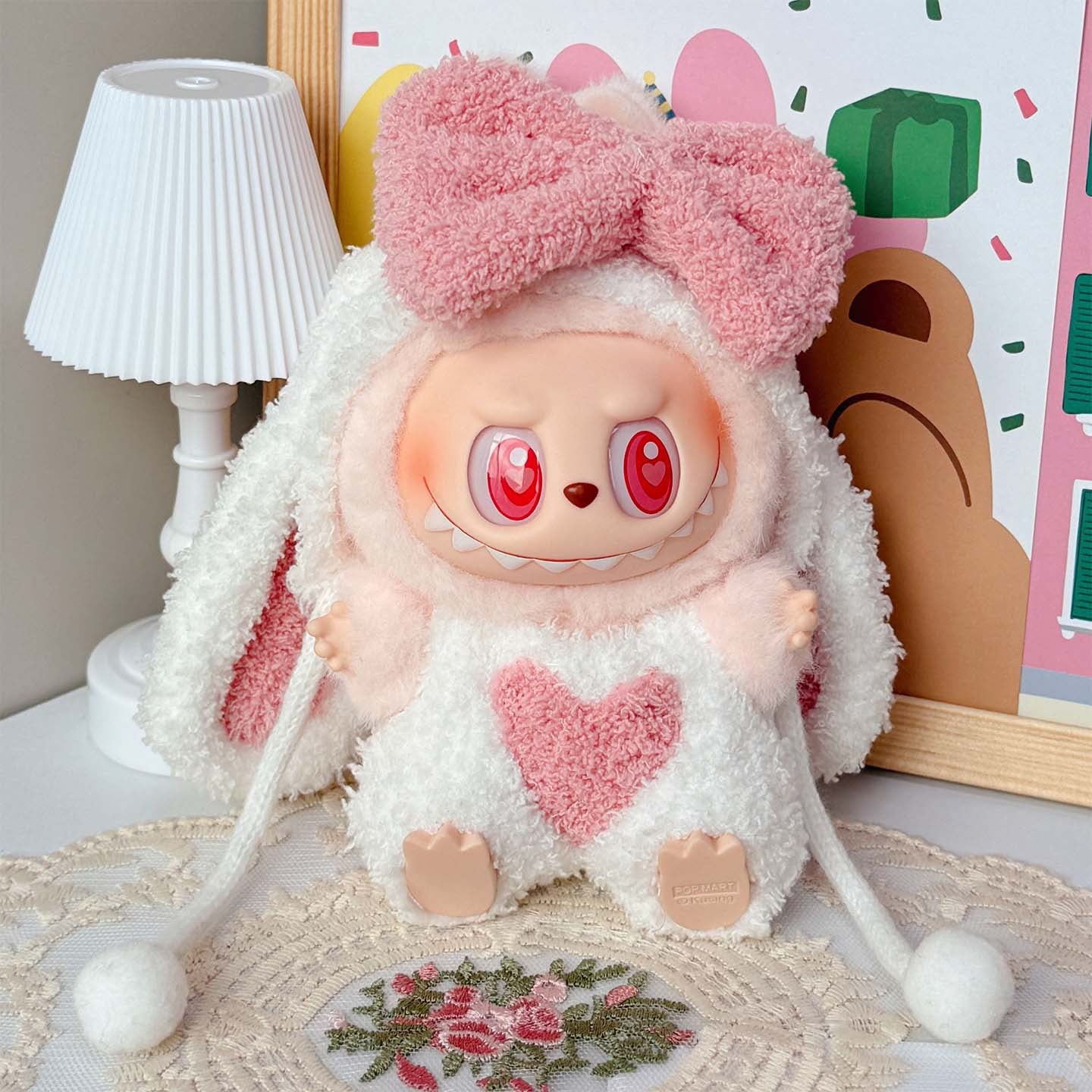 A ZoDoll Labubu Pink White Fluffy Bunny Onesie Plush Doll, featuring a pink bow, red eyes, and a heart on its chest, sits before a lamp and cartoon-themed background.