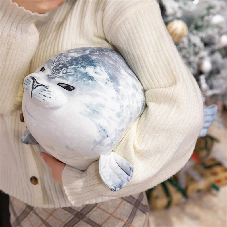 A person in a cream sweater holds the ZoDoll Seal Plush Toys Cute Chubby Seal Plush Pillow Soft Stuffed Seal Animal near a decorated Christmas tree with wrapped gifts underneath.