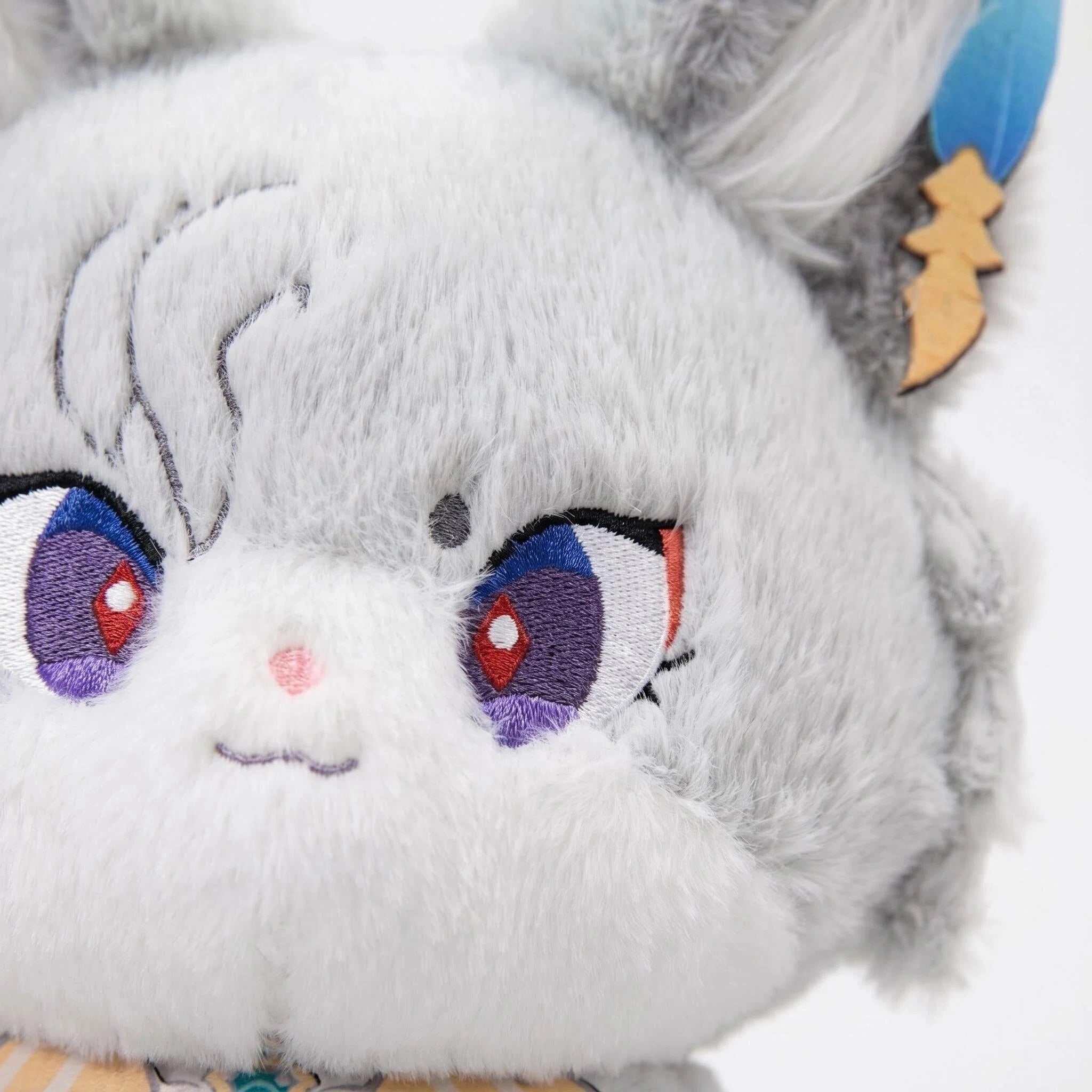 A close-up of a gray plush toy with big purple and red eyes, a small pink nose, and a subtle smile. The toy has fluffy fur, pointed ears, and wears a blue and gold accessory on one ear.