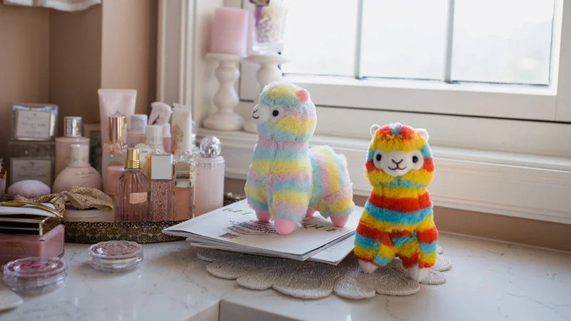 Two colorful plush alpaca toys sit on a white vanity next to makeup items, perfume bottles, and a window with soft daylight coming in. The vanity has a neat, pastel aesthetic.