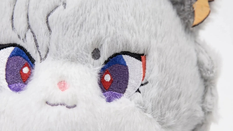 A close-up of a gray fox plush toy with big purple and red eyes, a small pink nose, and a subtle smile. The toy has fluffy fur, pointed ears, and wears a blue and gold accessory on one ear.
