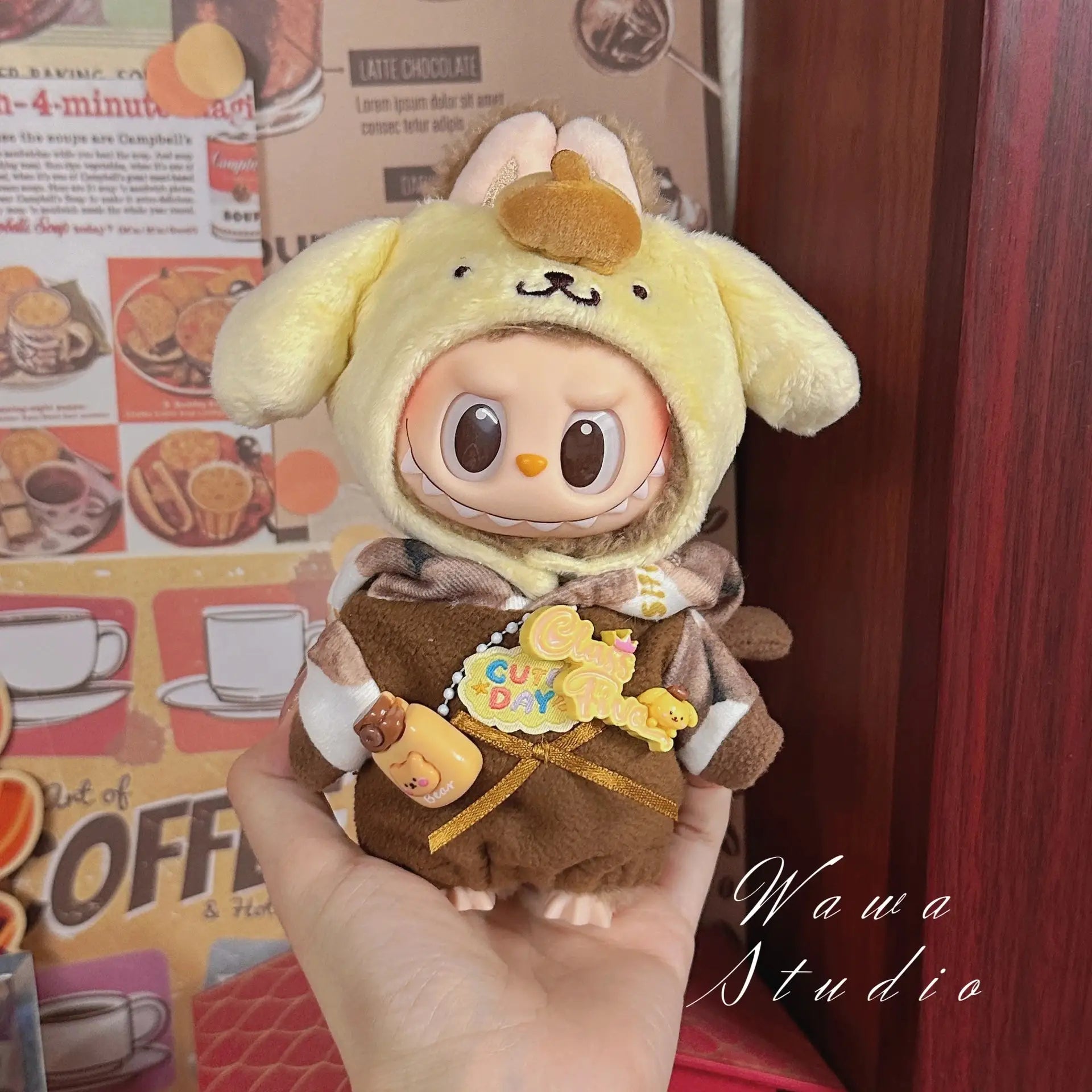 A hand holds a ZoDoll Labubu Sanrio Doll Clothes Set – Hello Kitty Style Outfit Collection, featuring a kawaii doll in a yellow animal hoodie and brown outfit with a Cute Day badge, displayed against a cafe-themed background.