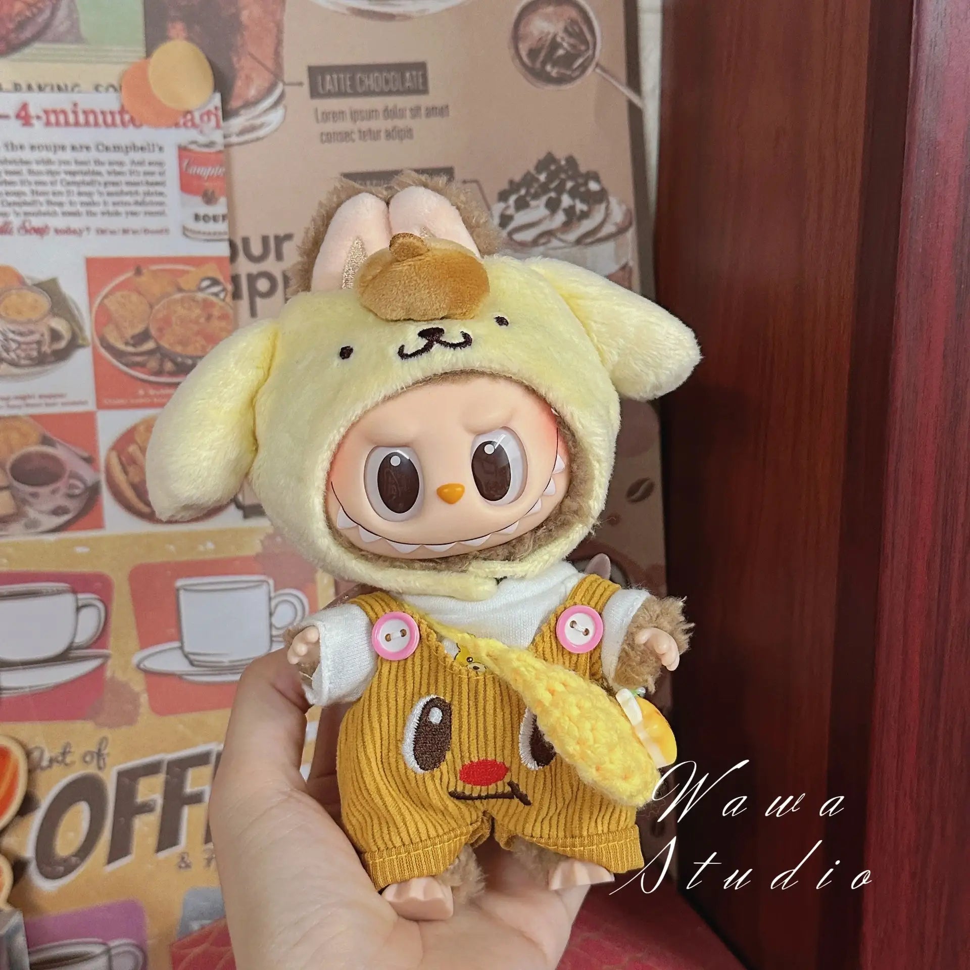 A ZoDoll Labubu Sanrio Doll in a Hello Kitty style outfit, featuring a yellow animal costume with floppy ears and brown overalls, is held before a wall adorned with food-themed prints.