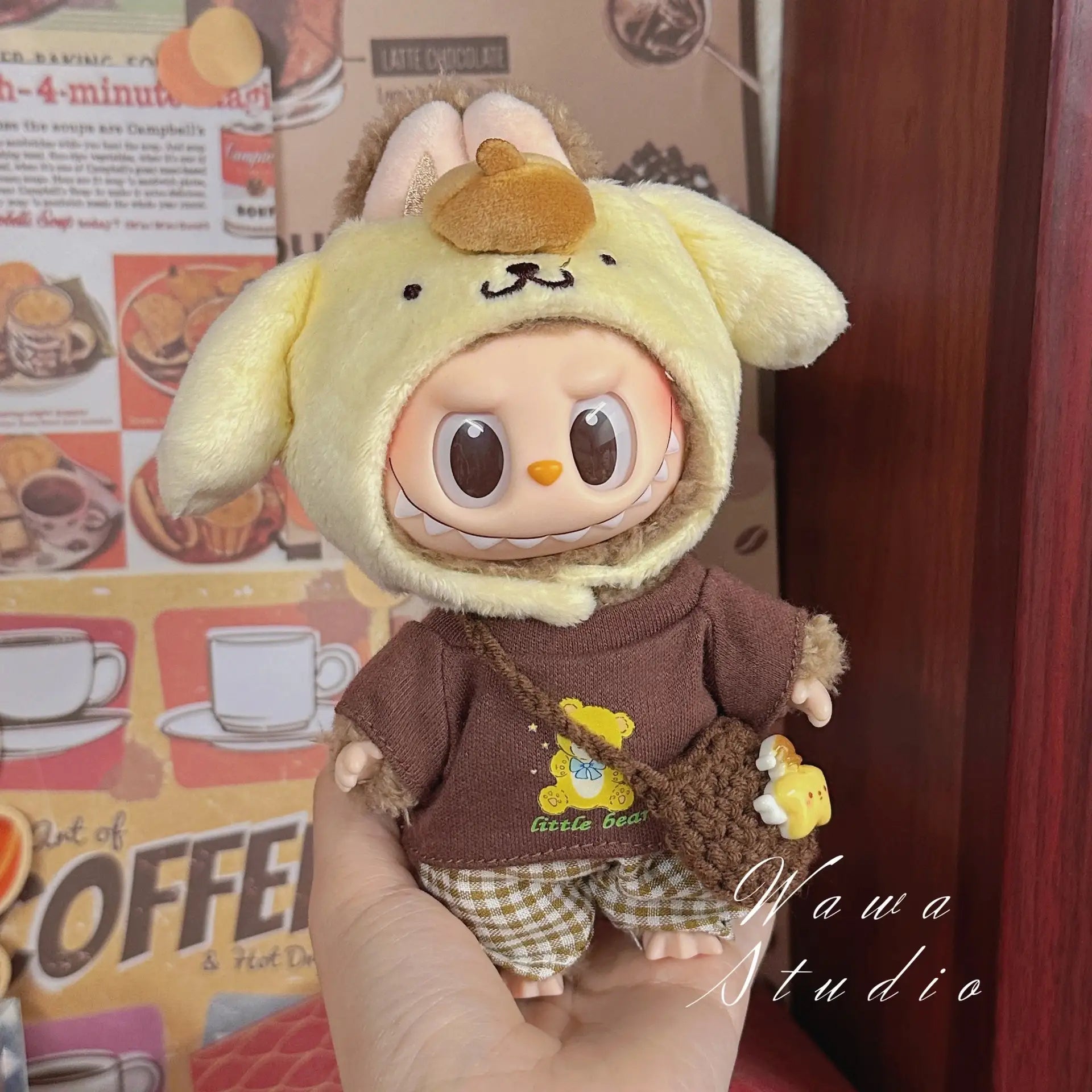 A ZoDoll Labubu Sanrio Doll Clothes Set in Hello Kitty style features a doll wearing a yellow animal-themed hood, brown shirt, plaid shorts, and small bag, posed in front of a coffee-themed background.