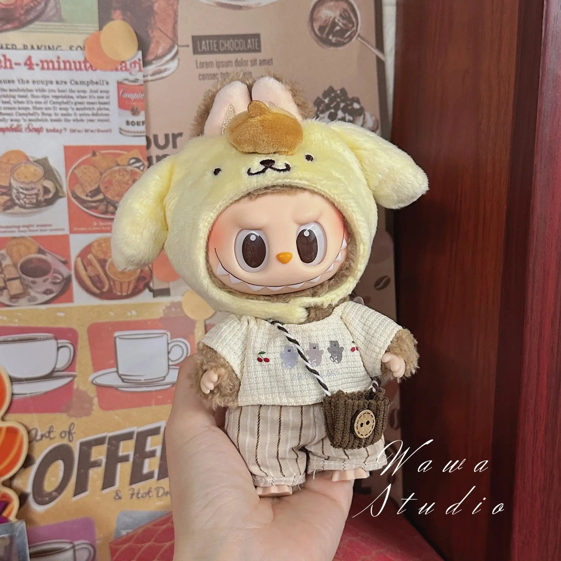 A hand holds a kawaii doll in yellow animal-themed clothes, set against a café backdrop with "Mawa Studio" in the corner—featuring the ZoDoll Labubu Sanrio Doll Clothes Set: Hello Kitty Style Outfit Collection.