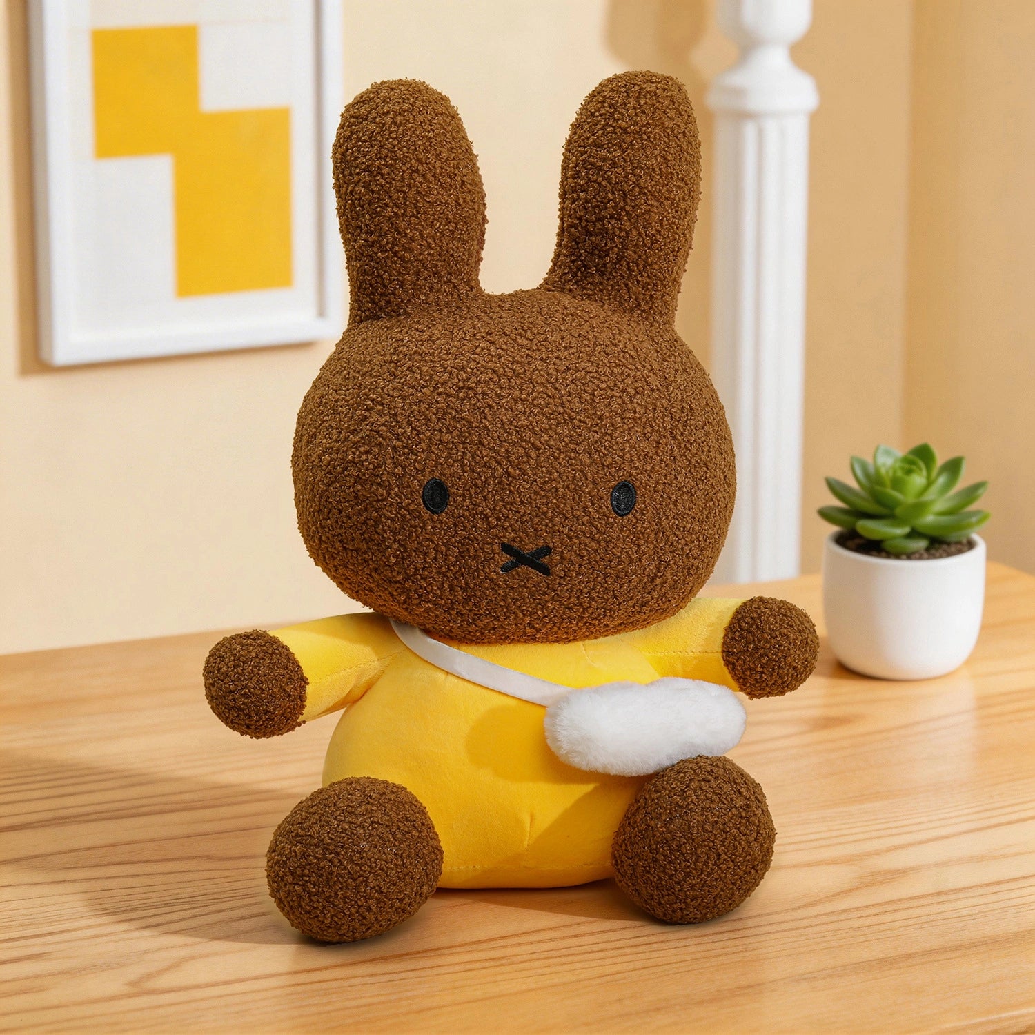 A ZoDoll Miffy Plush Toy—a brown bunny in a yellow outfit with a white bag—sits on a wooden table, with a potted succulent and framed art in the background. Available in 2 colors, sizes 30cm to 80cm.