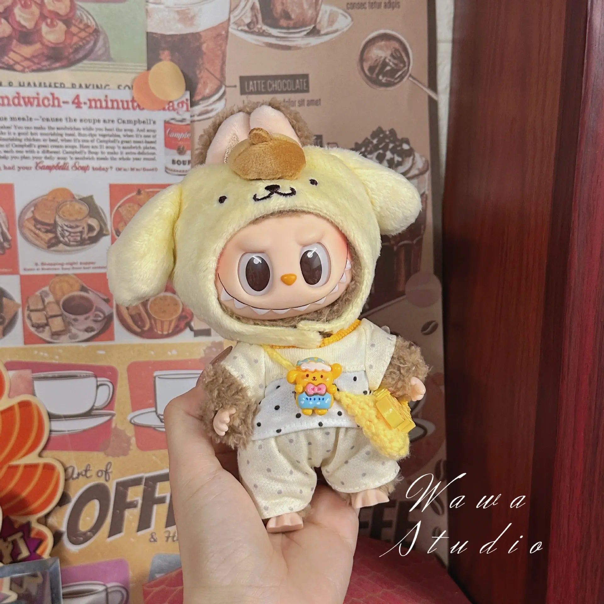 A hand holds a ZoDoll Labubu Sanrio Doll Clothes Set – Hello Kitty Style Outfit Collection doll in a yellow and brown animal costume with floppy ears, shown in front of a retro food and coffee poster. "Wawa Studio" appears at the bottom right.