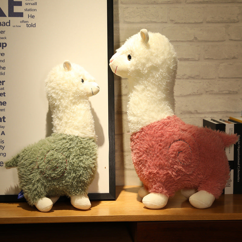 Two Cute Alpaca Plush Dolls by ZoDoll—one green and one pink, each 26cm—stand on a wooden surface against a white brick wall. The perfect soft plush toy for alpaca lovers to add to their collection.