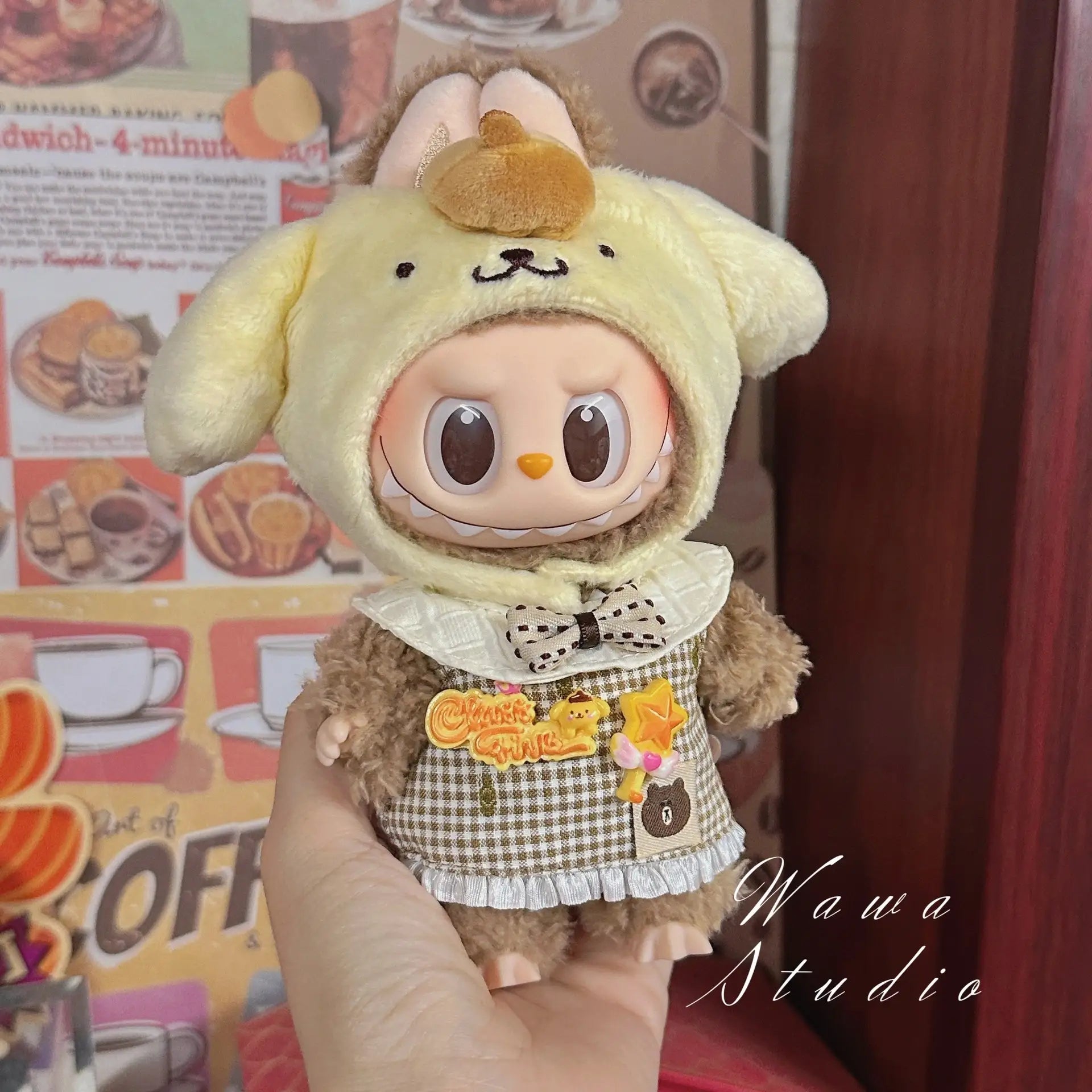 A hand holds a ZoDoll Labubu Sanrio Doll dressed in the Hello Kitty Style Outfit Collection, featuring a yellow hood and brown outfit with bow and patches, set against a colorful, food-themed background.
