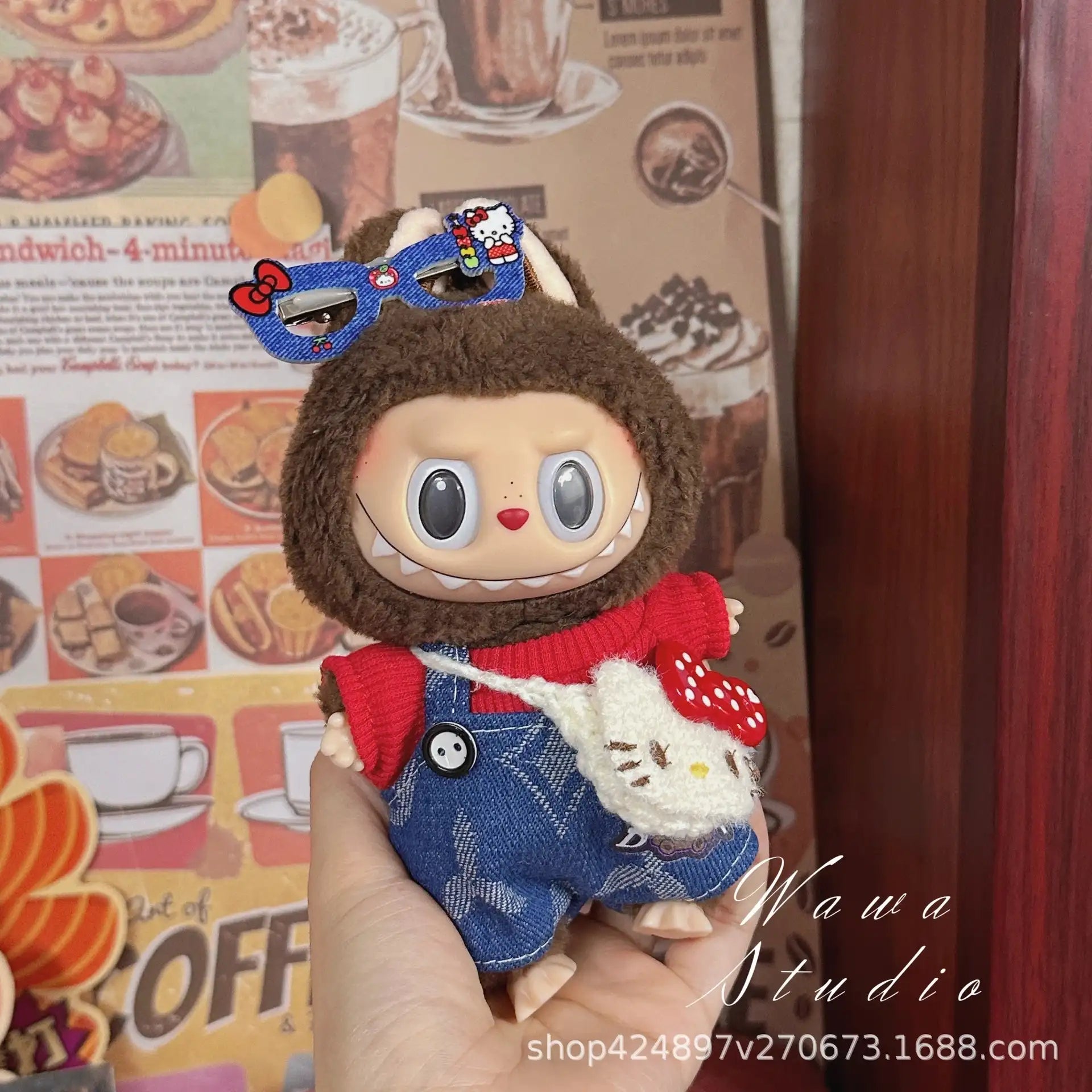 A hand holds a doll dressed in ZoDoll's Labubu Sanrio Doll Clothes Set – Hello Kitty Style Outfit Collection, featuring a brown animal hood, blue overalls, red shirt, and blue glasses. Coffee-themed posters and a white plush are in the background.