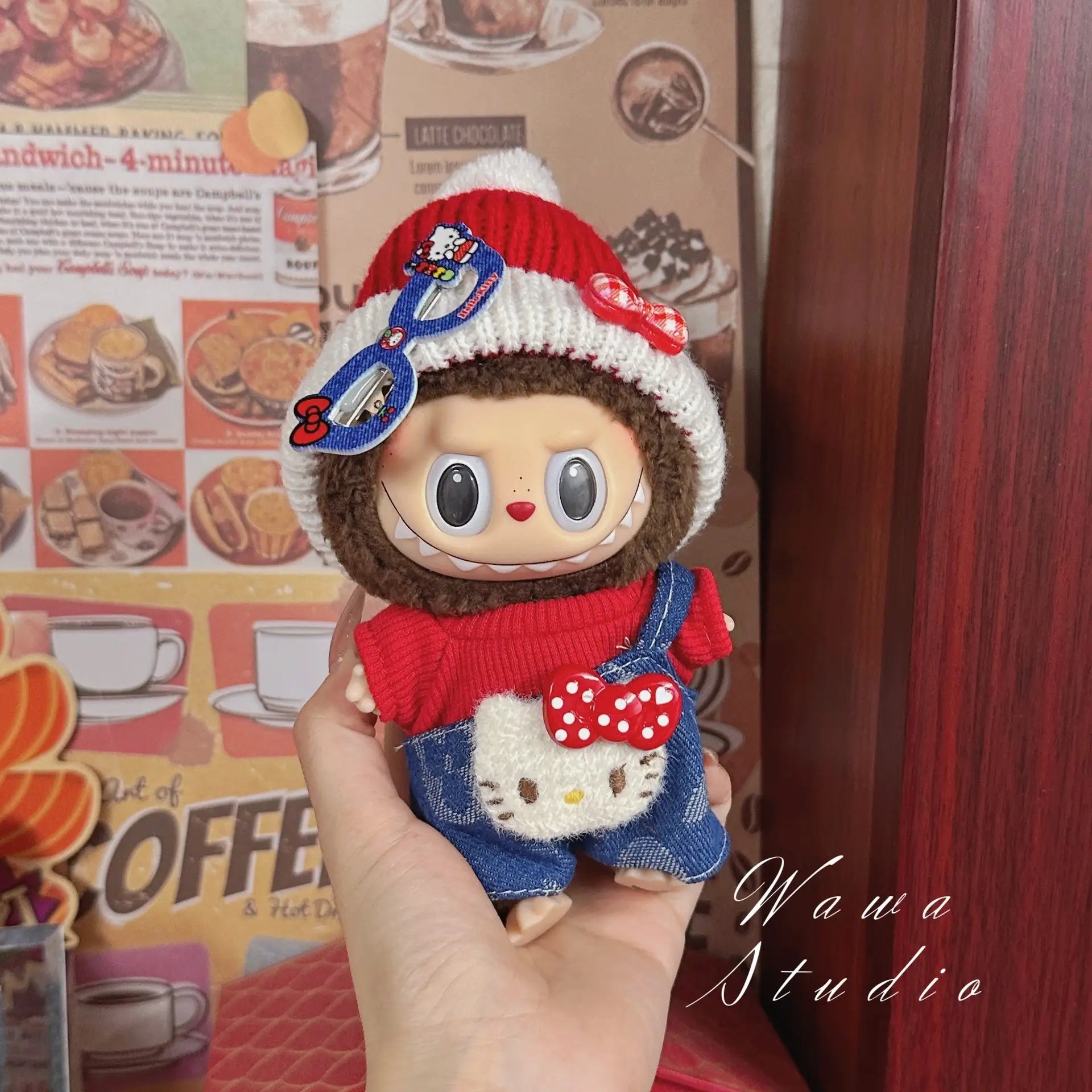 A hand holds a ZoDoll Labubu Sanrio Doll Clothes Set – Hello Kitty Style Outfit, featuring a red and white knit hat and denim overalls. Retro café-themed wallpaper decorates the background.