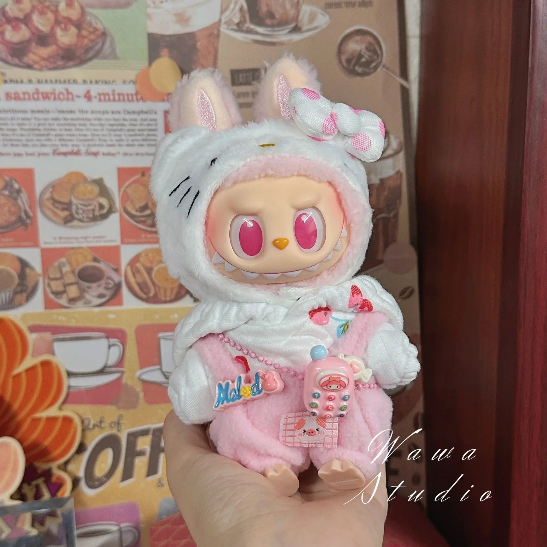 A person holds a ZoDoll Labubu Sanrio Doll in the Hello Kitty Style Outfit Collection, featuring a white bunny hat and pink clothes with a pacifier and necklace, in front of a food-themed poster.