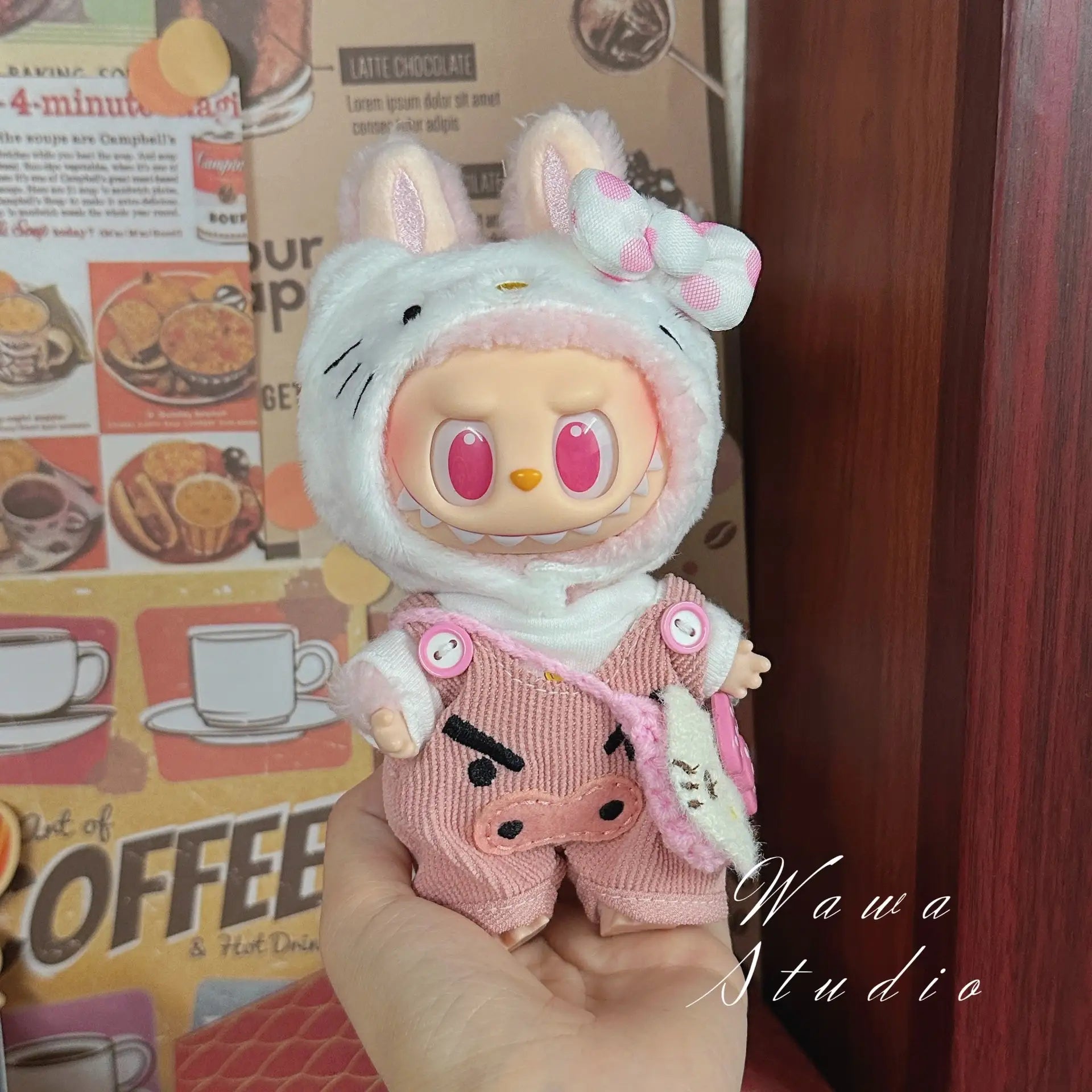 A hand holds a ZoDoll Labubu Sanrio Doll Clothes Set – Hello Kitty Style Outfit, featuring a white hood with pink ears and pig-themed details. A coffee-themed wall poster is visible in the background.