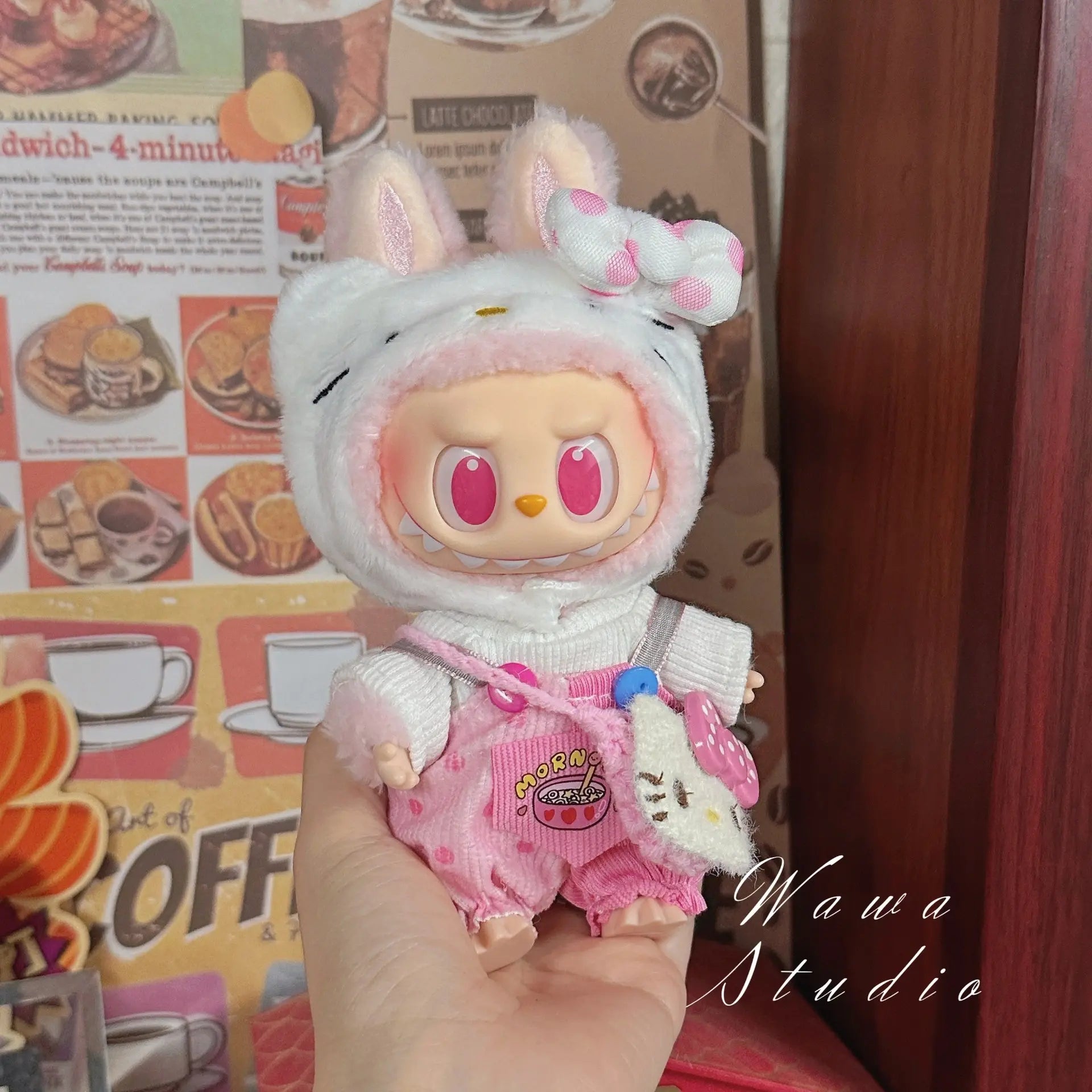 A person holds a ZoDoll Labubu Sanrio Doll Clothes Set—Hello Kitty Style Outfit Collection—featuring a kawaii hoodie with bunny ears and overalls, against a backdrop with food posters.