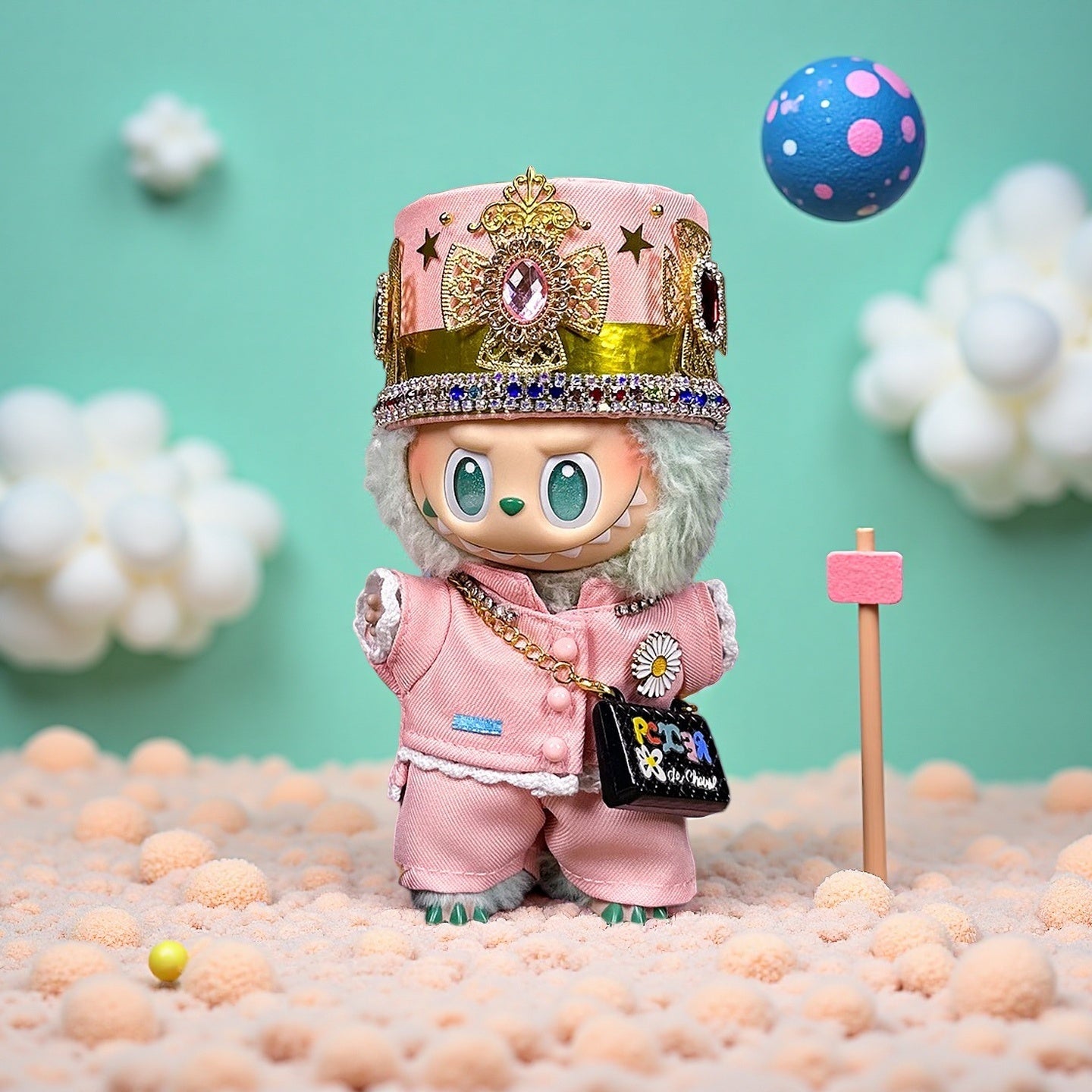 The ZoDoll Labubu Pink Royal Twill Suit Set with Crown Hat and Mini Bag stands on a textured surface with a pink sign, set against a teal background featuring floating clouds and a patterned ball.