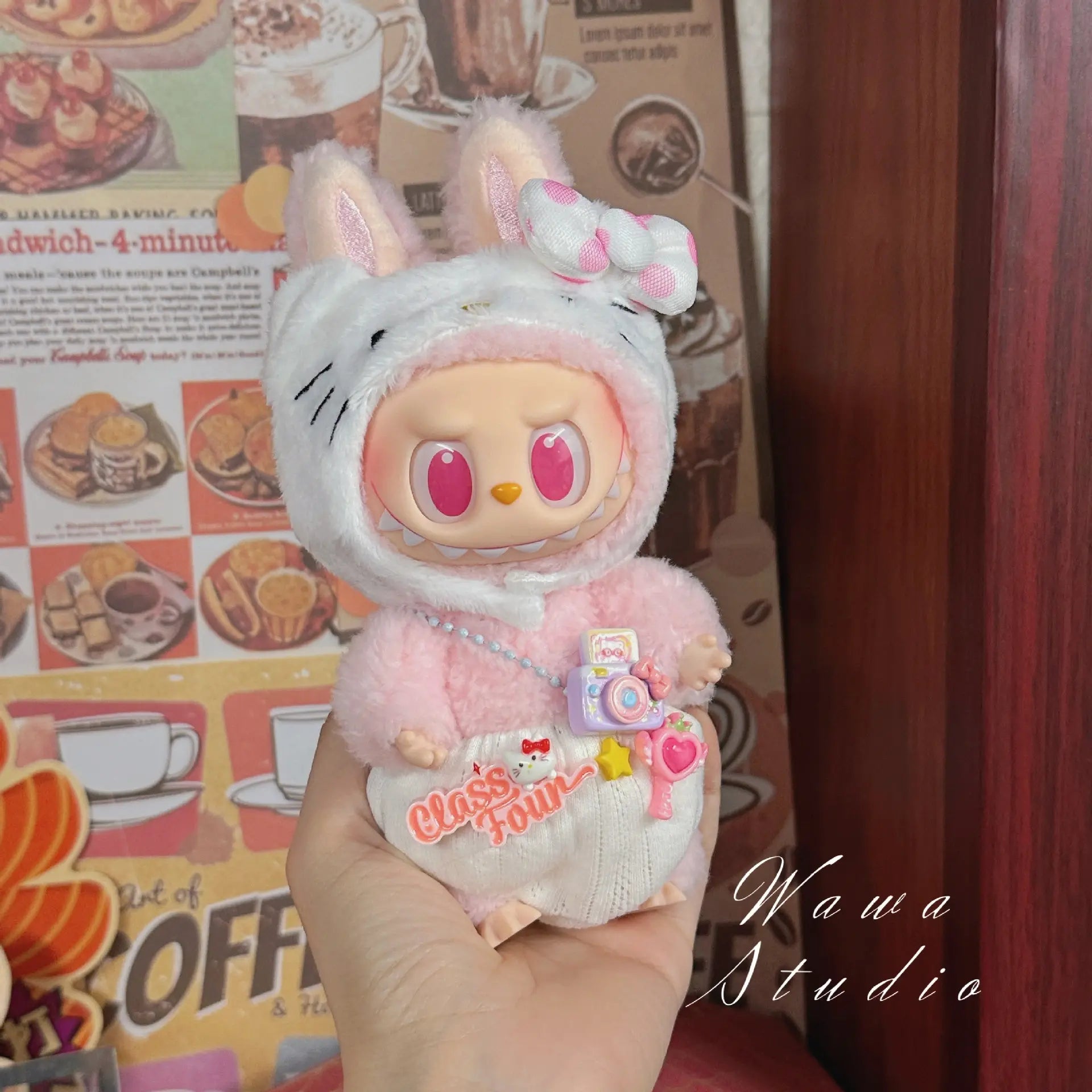 A hand holds a ZoDoll Labubu Sanrio Doll in a Hello Kitty style outfit from the Doll Clothes Set, complete with a camera accessory, against a retro café-themed background.