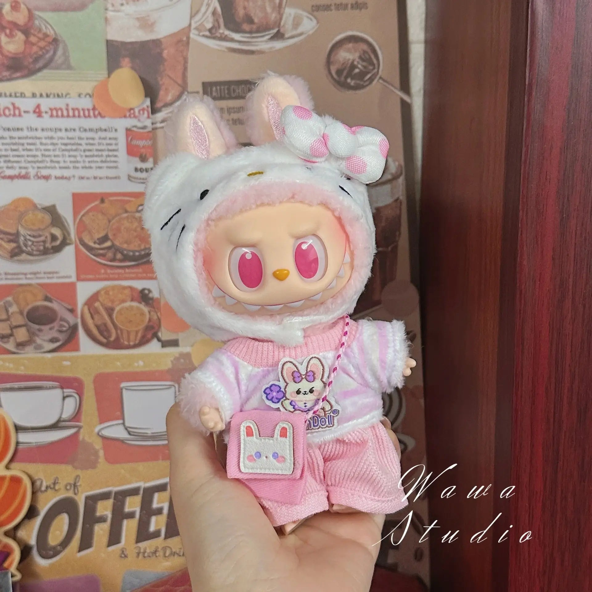 A hand holds a ZoDoll Labubu Sanrio Doll Clothes Set – Hello Kitty Style Outfit Collection, with the doll in a pink and white kawaii bunny outfit, shown against a coffee-themed background.