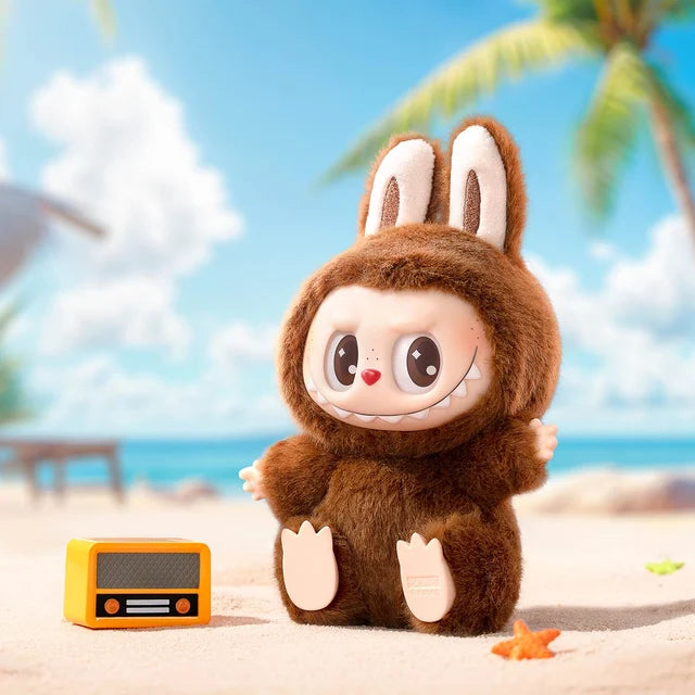 A smiling Labubu “Duoduo” Plush Keychain – Have a Seat Series Brown Freckled Bunny by POP MART stands on a sandy beach near a small yellow radio, with palm trees and blue sky in the background—part of The Monsters.