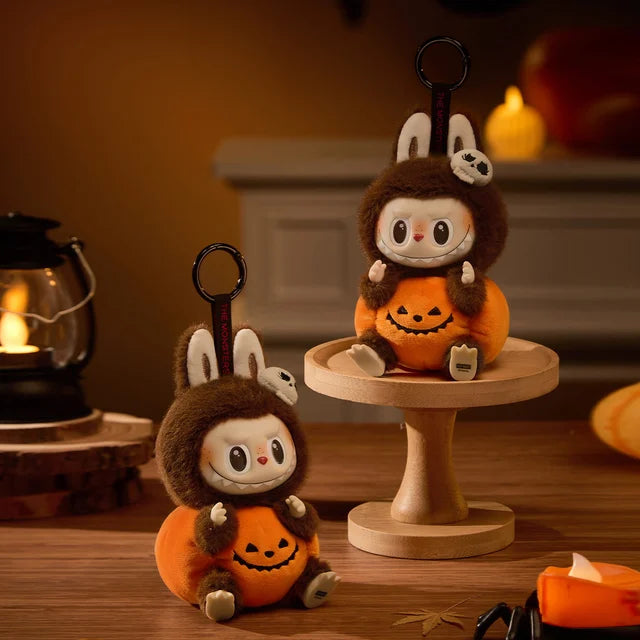 Two POP MART THE MONSTERS – Happy Halloween Party Labubu 15cm Vinyl Plush Keychains (Pumpkin Edition) featuring rabbit characters in brown hoodies atop pumpkin bases are displayed with autumn décor—ideal Halloween collectibles.