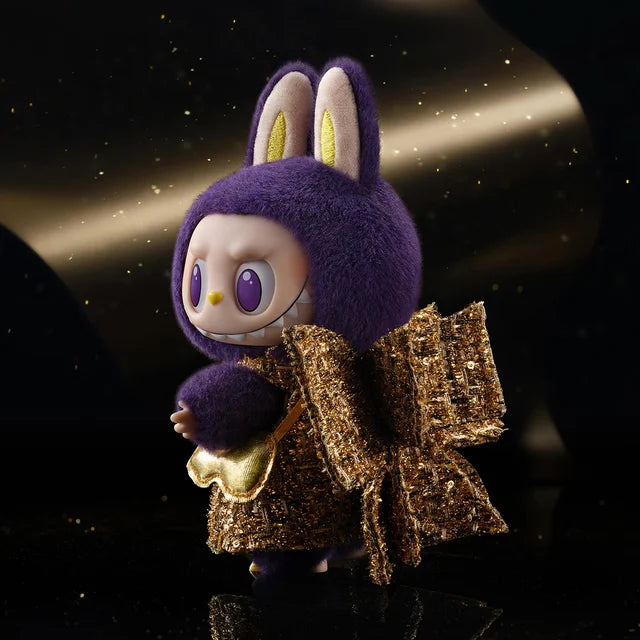 The Labubu × PRONOUNCE Wings of Fortune – Limited Edition Designer Plush Vinyl Toy by POP MART features a fuzzy purple bunny suit with gold ears, large eyes, a wide grin, and a gold bow against a dark, sparkling backdrop.