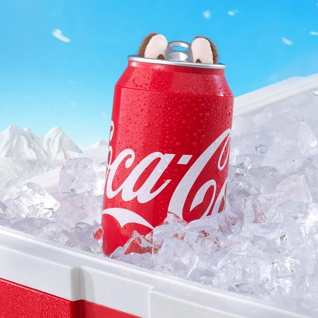 A POP MART Labubu Secret Edition “Mysterious Guest – Coca‑Cola” Plush Keychain collectible, with bear ears and red can design, sits upright in an ice-filled cooler, mountains visible in the background.