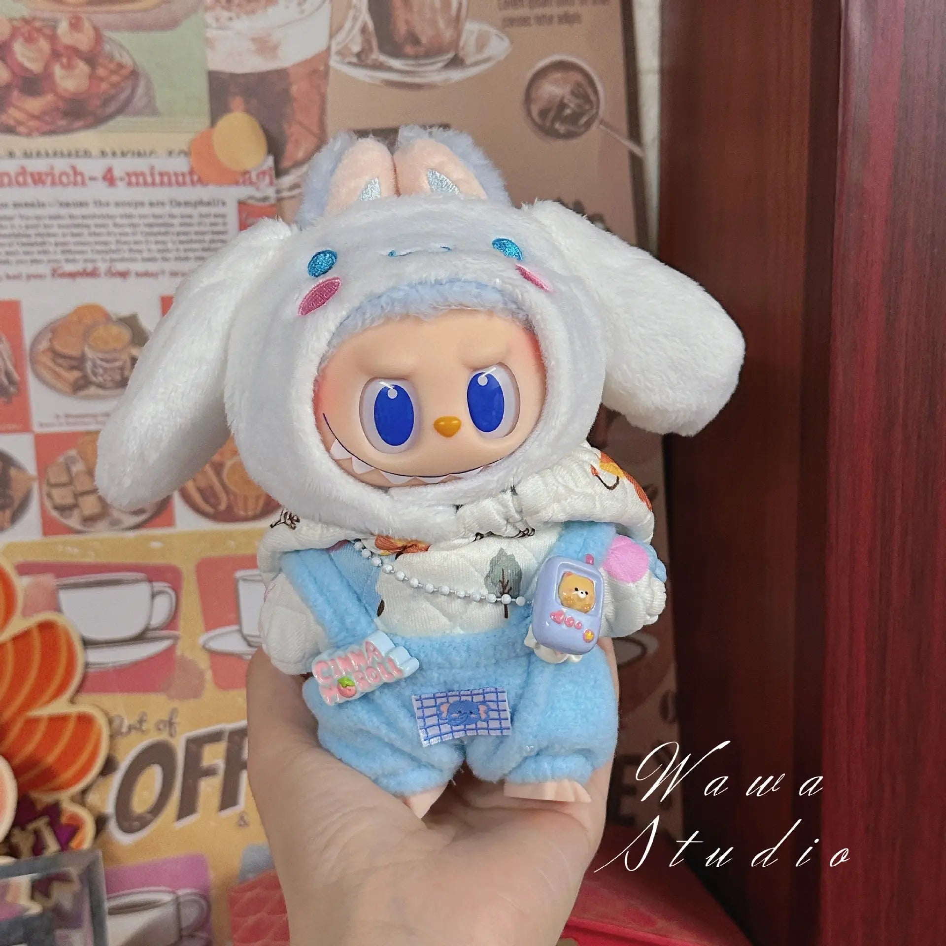 A hand displays the ZoDoll Labubu Sanrio Doll Clothes Set – Hello Kitty Style Outfit Collection, featuring a doll in a white bunny hoodie, blue overalls, and patterned scarf with kawaii fashion, set against a café-themed background.