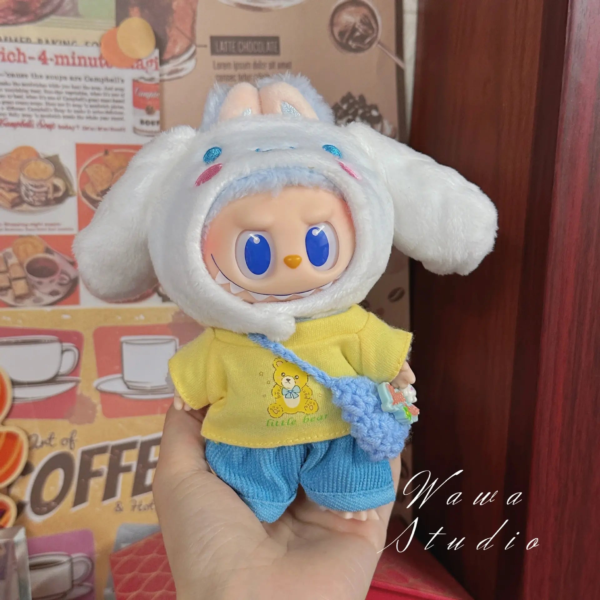 A hand holds a ZoDoll Labubu Sanrio Doll dressed in the Hello Kitty Style Outfit Collection, featuring a white bunny hat, yellow bear-print shirt, blue shorts, and crocheted bag against a coffee-themed wall.