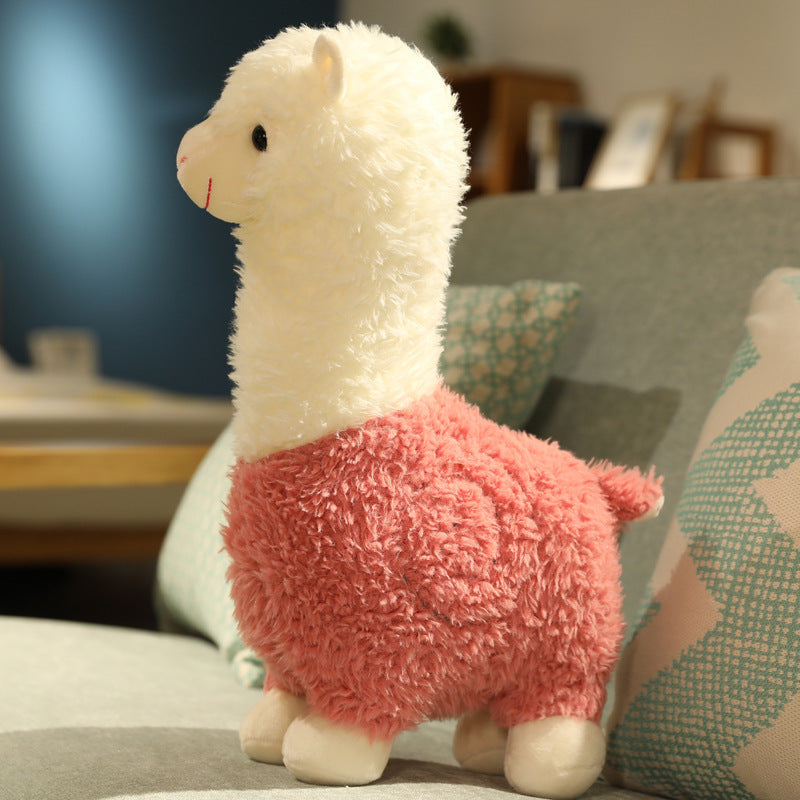 The ZoDoll Cute Alpaca Plush Doll - Soft Alpaca Plush Toy 26cm features a white head and neck with a pink body, sitting upright on a gray sofa next to patterned pillows.
