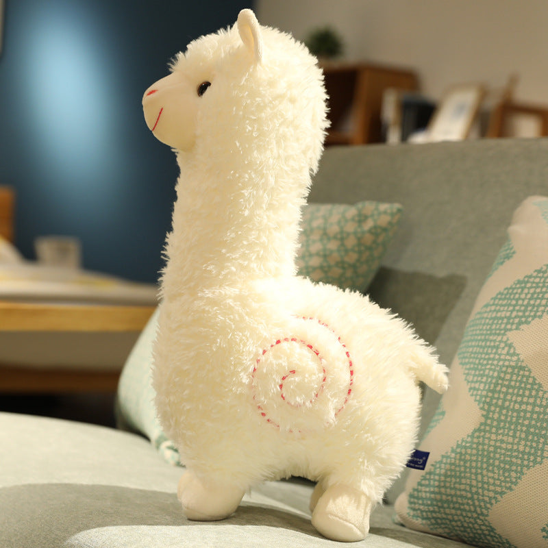 The ZoDoll Cute Alpaca Plush Doll, a 26cm soft alpaca plush toy featuring a spiral design, rests on a light-colored sofa with patterned cushions in the background.