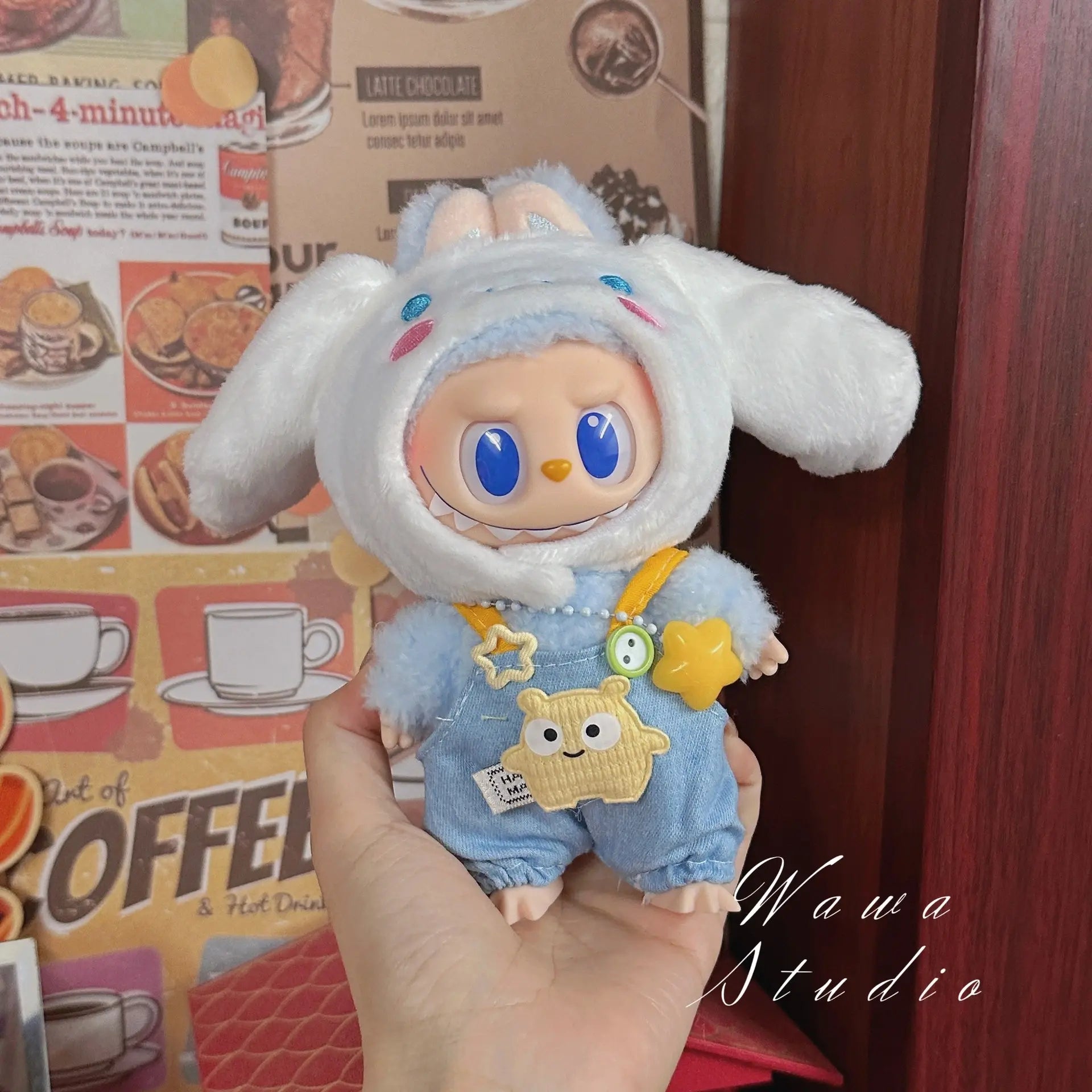 A person holds a ZoDoll Labubu Sanrio Doll dressed in the Hello Kitty Style Outfit Collection, featuring a fluffy bunny hat and blue overalls, standing before a coffee-themed wall.