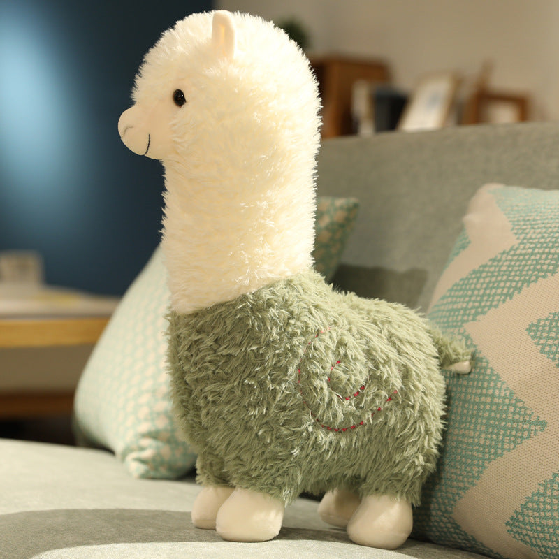 The ZoDoll Cute Alpaca Plush Doll, a 26cm soft alpaca plush toy with a white head and green body, sits upright on a couch with patterned pillows in the background.