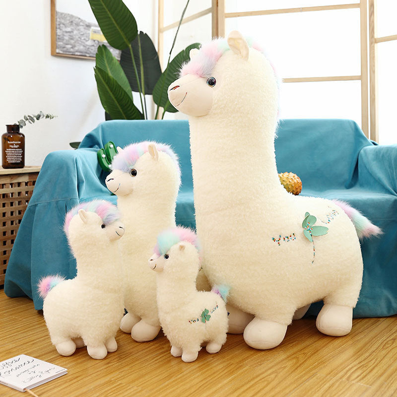 Four ZoDoll Cute Alpaca Plush Toy - Soft Stuffed Alpaca Plush Dolls with pastel manes are arranged on a wooden floor in front of a blue sofa, surrounded by plants and a window in a bright, cheerful room.