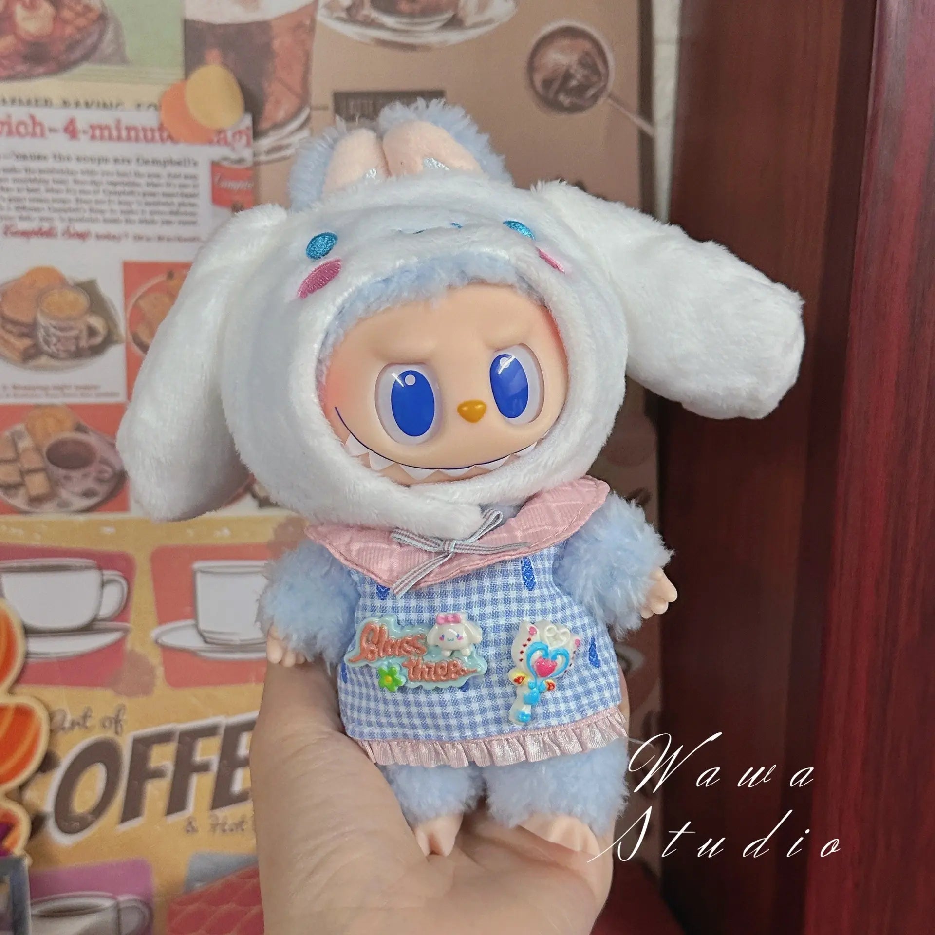 A hand holds a ZoDoll Labubu Sanrio Doll Clothes Set – Hello Kitty Style Outfit Collection, featuring a cute blue and white bunny costume with long ears, embroidered accents, and a pink collar. Food-themed posters add extra charm to this playful look.