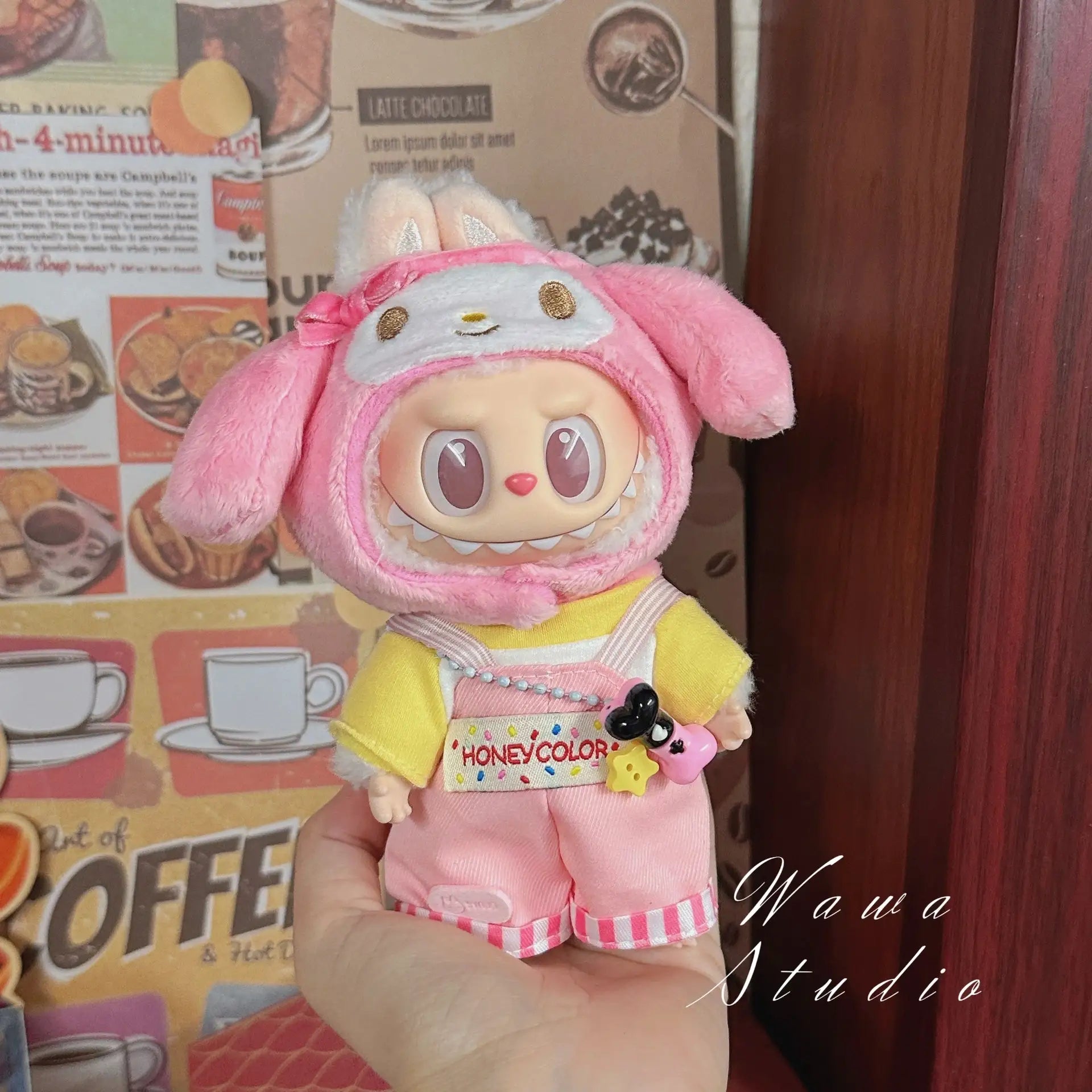 A hand holds a ZoDoll Labubu Sanrio Doll in a Hello Kitty style outfit from the Doll Clothes Set, featuring kawaii fashion against a coffee-themed background.