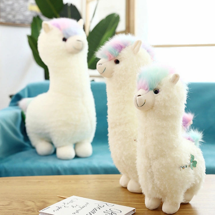Three ZoDoll Cute Alpaca Plush Toy - Soft Stuffed Alpaca Plush Dolls with pastel manes and tails are displayed on a wooden table and a blue sofa, set against a backdrop of leafy green plants.